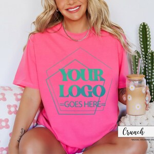 May include: A vibrant pink t-shirt with the text "YOUR LOGO GOES HERE" in teal. The shirt is paired with a daisy-decorated glass and a straw. The word "Crunch" is displayed in a white rectangle.
