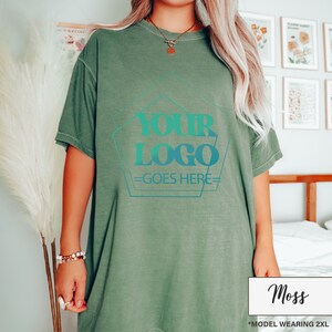 May include: A moss green t-shirt with a teal and blue logo design that reads "YOUR LOGO GOES HERE". The shirt is a relaxed fit and has short sleeves. The model is wearing a 2XL size.