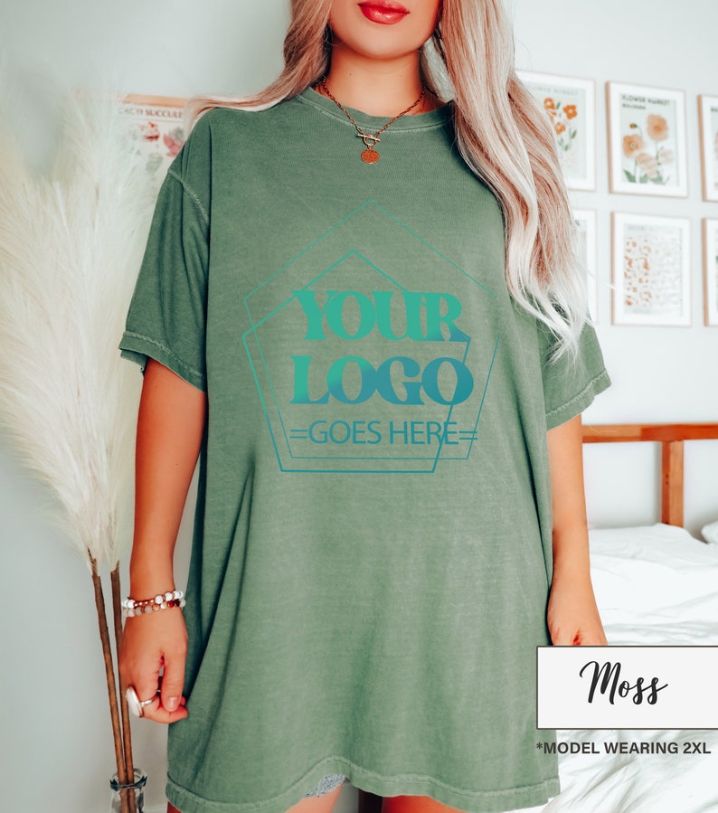 May include: A moss green t-shirt with a teal and blue logo design that reads "YOUR LOGO GOES HERE". The shirt is a relaxed fit and has short sleeves. The model is wearing a 2XL size.