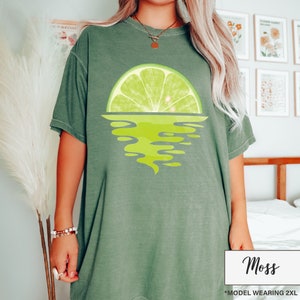 May include: A green oversized t-shirt with a lime wedge graphic. The lime wedge is half submerged in water, creating a sunset effect. The text "Moss" is printed on the bottom right corner of the image. The text "*MODEL WEARING 2XL" is printed below the "Moss" text.