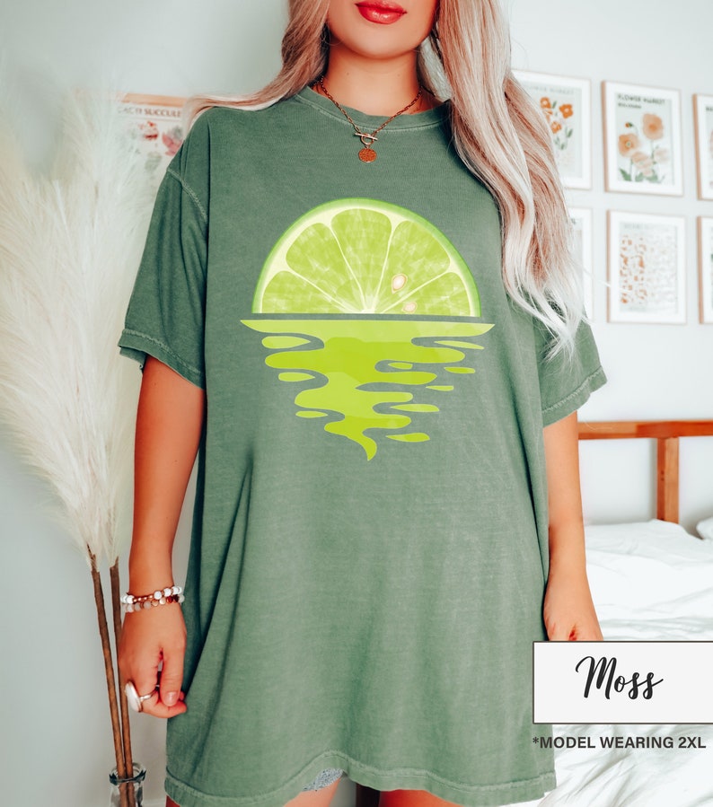 May include: A green oversized t-shirt with a lime wedge graphic. The lime wedge is half submerged in water, creating a sunset effect. The text "Moss" is printed on the bottom right corner of the image. The text "*MODEL WEARING 2XL" is printed below the "Moss" text.