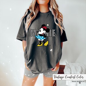 May include: Dark gray t-shirt featuring a Minnie Mouse graphic. The graphic includes the name "MINNIE" in a checkered pattern, and Minnie is wearing a blue polka dot dress and yellow shoes. The shirt is labeled "Vintage Comfort Color".