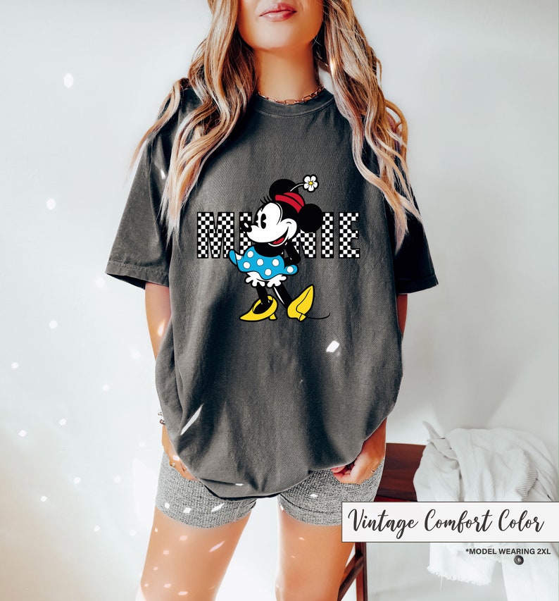 May include: Dark gray t-shirt featuring a Minnie Mouse graphic. The graphic includes the name "MINNIE" in a checkered pattern, and Minnie is wearing a blue polka dot dress and yellow shoes. The shirt is labeled "Vintage Comfort Color".