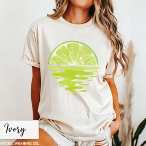 May include: A woman is wearing a light beige t-shirt with a lime wedge graphic. The lime wedge is reflected in the water below it. The text "Ivory" is at the bottom of the image.