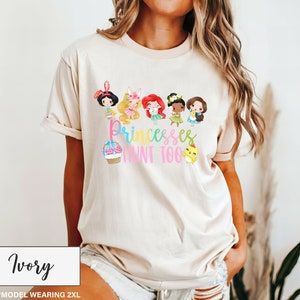 May include: Ivory-colored t-shirt featuring a colorful graphic of cartoon princesses with the text "Princesses Hunt Too." The design includes a cupcake and a chick. The shirt is a relaxed fit.