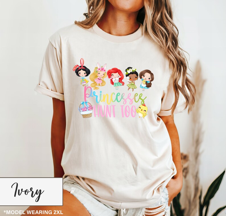 May include: Ivory-colored t-shirt featuring a colorful graphic of cartoon princesses with the text "Princesses Hunt Too." The design includes a cupcake and a chick. The shirt is a relaxed fit.