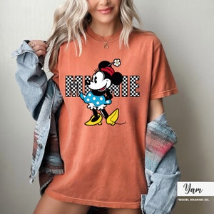 May include: Orange-colored t-shirt featuring a graphic of Minnie Mouse. Minnie is wearing a blue polka-dot dress, yellow shoes, and a red hat with a flower. The word "MINNIE" is printed in a checkered pattern.
