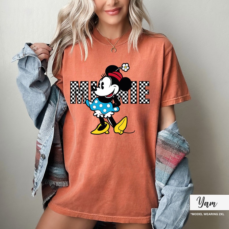 May include: Orange-colored t-shirt featuring a graphic of Minnie Mouse. Minnie is wearing a blue polka-dot dress, yellow shoes, and a red hat with a flower. The word "MINNIE" is printed in a checkered pattern.