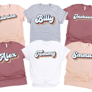 May include: Six t-shirts with different names printed in a retro rainbow font. The names are Sabrina, Billy, Jackson, Alex, Tommy, and Emma.