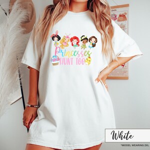 May include: White t-shirt featuring a colorful graphic of cartoon princesses and the text "Princesses Hunt Too." The design includes an Easter basket, chicks, and various princess characters. The shirt is a relaxed fit.