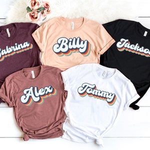 May include: Five t-shirts with different names printed on them in a retro rainbow font. The names are Sabrina, Billy, Jackson, Alex, and Tommy.