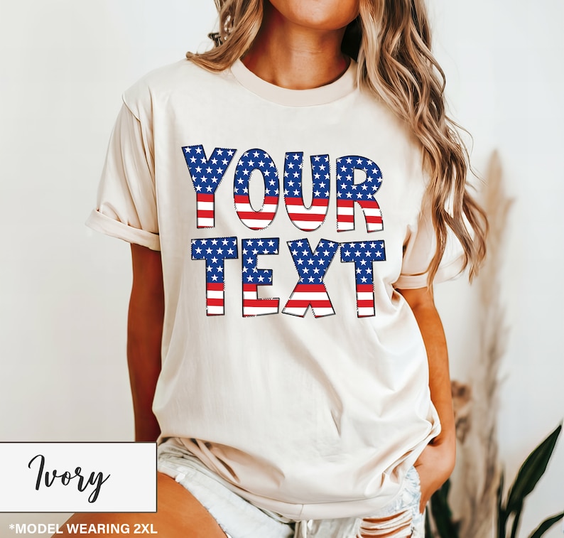 May include: Ivory-colored t-shirt with the text "YOUR TEXT" in a patriotic design of red and white stripes and blue stars. The shirt is a relaxed fit and has short sleeves. The text is in a bold, blocky font.