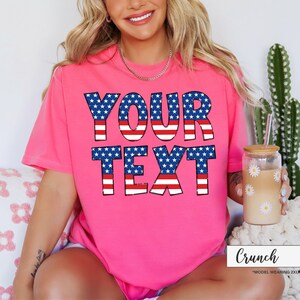 May include: A bright pink t-shirt with the words "YOUR TEXT" in a patriotic design of blue stars and red and white stripes. The shirt is short-sleeved and has a casual fit. A glass with a daisy design is held in the hand.