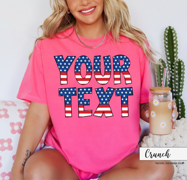 May include: A bright pink t-shirt with the words "YOUR TEXT" in a patriotic design of blue stars and red and white stripes. The shirt is short-sleeved and has a casual fit. A glass with a daisy design is held in the hand.