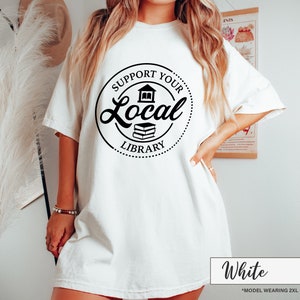 May include: White t-shirt with a black circular graphic that reads "Support Your Local Library." The graphic includes a house and two books. The shirt is a loose fit and has short sleeves. The text "White" is at the bottom.