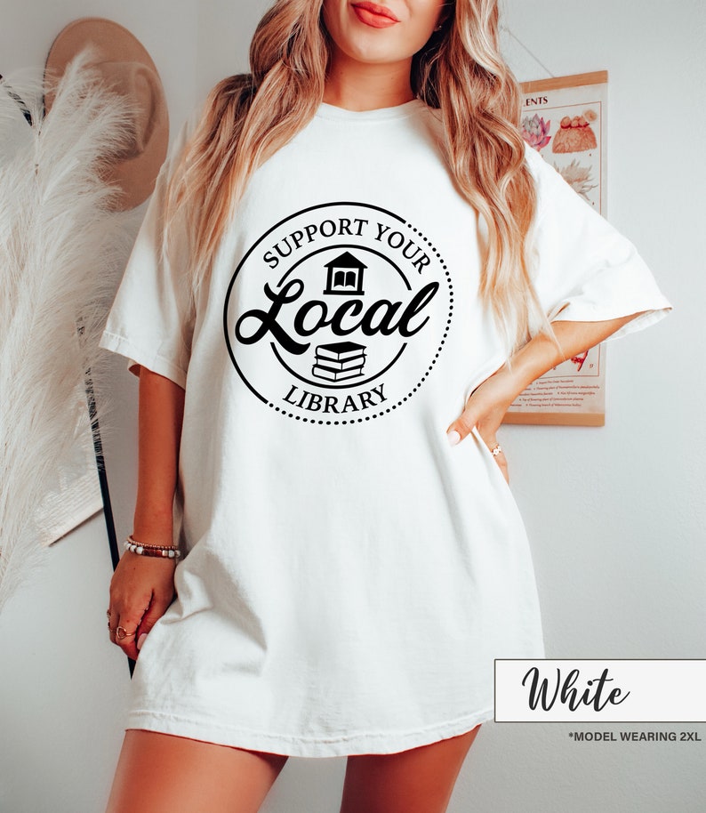 May include: White t-shirt with a black circular graphic that reads "Support Your Local Library." The graphic includes a house and two books. The shirt is a loose fit and has short sleeves. The text "White" is at the bottom.