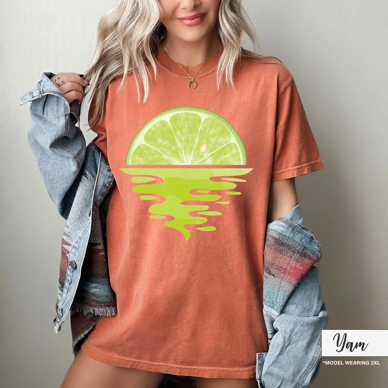 May include: A burnt orange t-shirt with a lime wedge graphic. The lime wedge is depicted as a sunset over a body of water. The model is wearing a size 2XL.