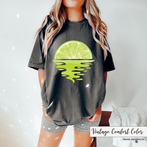 May include: A dark gray t-shirt with a lime wedge graphic. The lime wedge is half submerged in water, creating a sunset effect. The text "Vintage Comfort Color" is printed on the bottom of the image.
