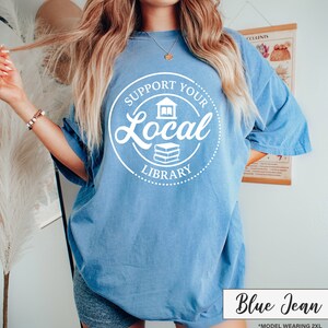 May include: A blue jean colored t-shirt with the text "Support Your Local Library" in a white circular design. The shirt has short sleeves and a relaxed fit. The text "Blue Jean" is in a white box at the bottom.