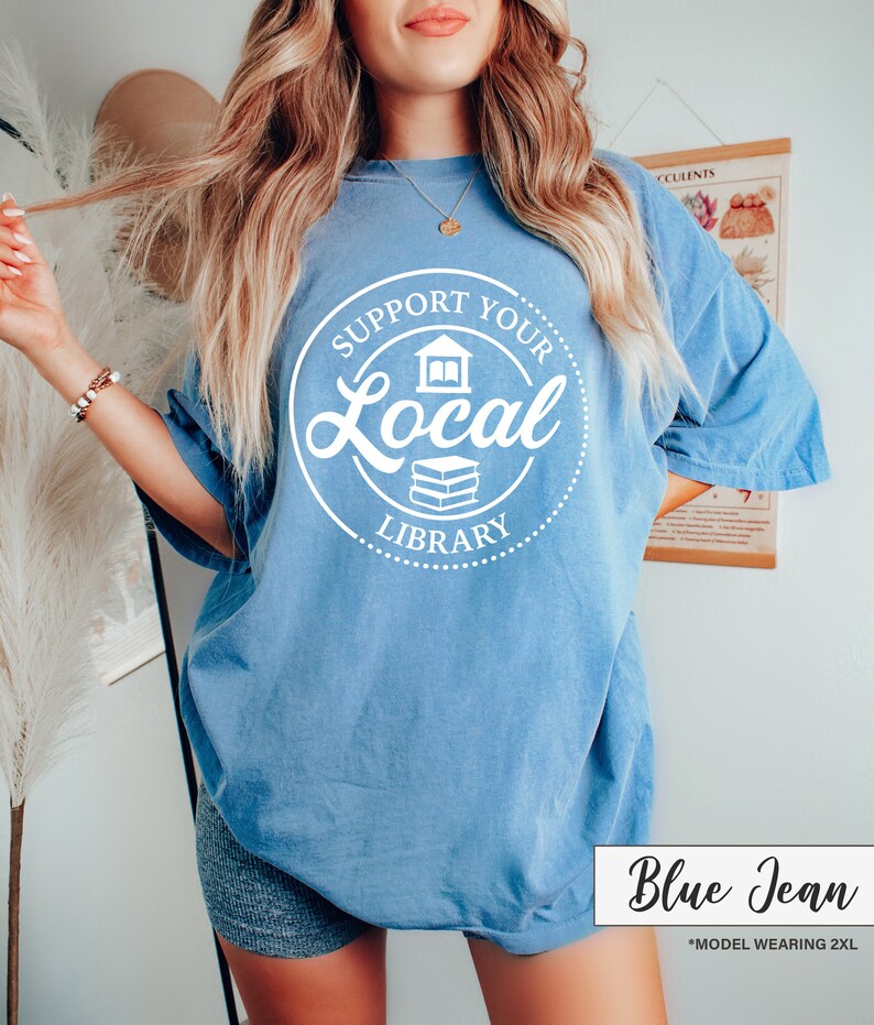 May include: A blue jean colored t-shirt with the text "Support Your Local Library" in a white circular design. The shirt has short sleeves and a relaxed fit. The text "Blue Jean" is in a white box at the bottom.