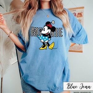 May include: Blue jean colored t-shirt featuring a Minnie Mouse graphic. The graphic includes the word "MINNIE" in a checkered pattern, and Minnie Mouse is wearing a polka dot dress and yellow shoes. The text "Blue Jean" is at the bottom.