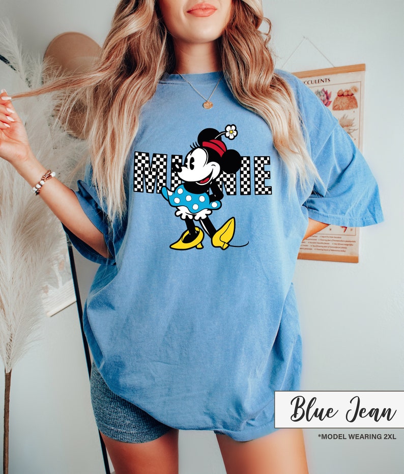 May include: Blue jean colored t-shirt featuring a Minnie Mouse graphic. The graphic includes the word "MINNIE" in a checkered pattern, and Minnie Mouse is wearing a polka dot dress and yellow shoes. The text "Blue Jean" is at the bottom.