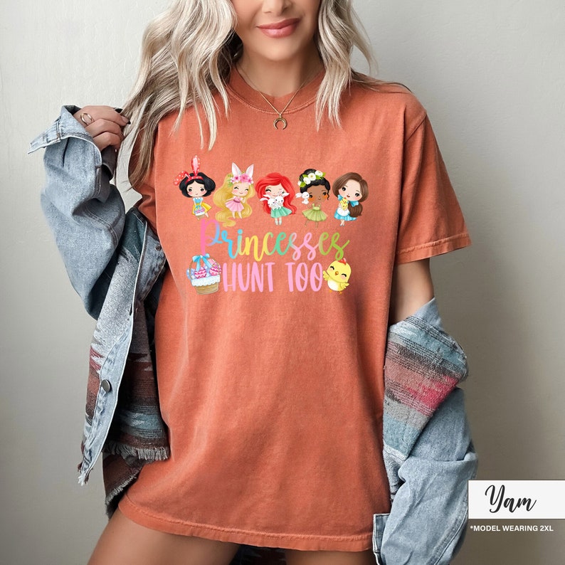May include: Burnt orange t-shirt featuring a graphic of cartoon princesses and the text "Princesses Hunt Too". The design includes a basket, a chick, and several princesses. The model is wearing a 2XL size.