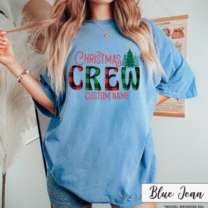 Może przedstawiać: Jasnoniebieska koszulka z napisem "Christmas Crew" w czerwono-zieloną kratę i zieloną grafiką choinki. Pod spodem znajduje się tekst "Custom Name". Koszulka jest oznaczona jako "Blue Jean".