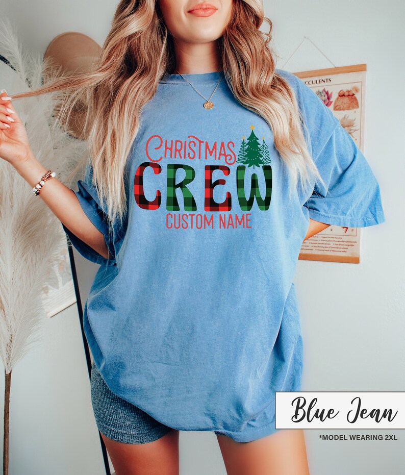 Może przedstawiać: Jasnoniebieska koszulka z napisem "Christmas Crew" w czerwono-zieloną kratę i zieloną grafiką choinki. Pod spodem znajduje się tekst "Custom Name". Koszulka jest oznaczona jako "Blue Jean".
