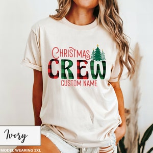 Może przedstawiać: Kremowa koszulka z napisem "Christmas Crew" w czerwone i zielone kratki. Koszulka posiada dwa zielone choinki i napis "Custom Name" w kolorze czerwonym. Modelka nosi rozmiar 2XL.