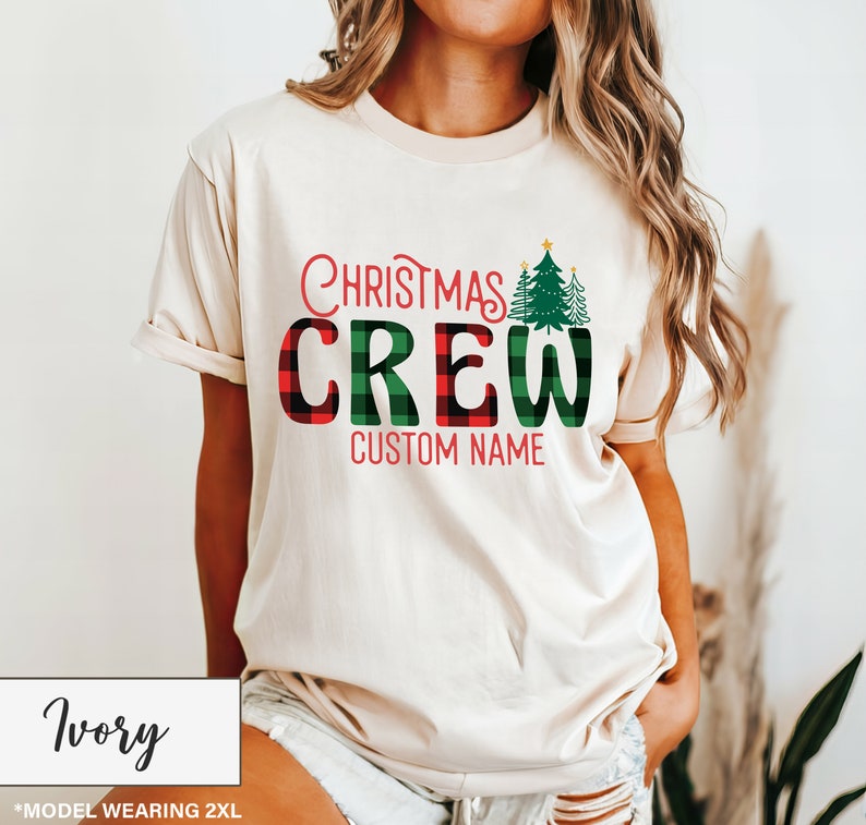 Może przedstawiać: Kremowa koszulka z napisem "Christmas Crew" w czerwone i zielone kratki. Koszulka posiada dwa zielone choinki i napis "Custom Name" w kolorze czerwonym. Modelka nosi rozmiar 2XL.