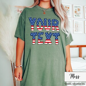 May include: Moss green t-shirt with the words "YOUR TEXT" in a patriotic design of red and white stripes and blue stars. The shirt is a loose fit and has short sleeves. The model is wearing a 2XL size.