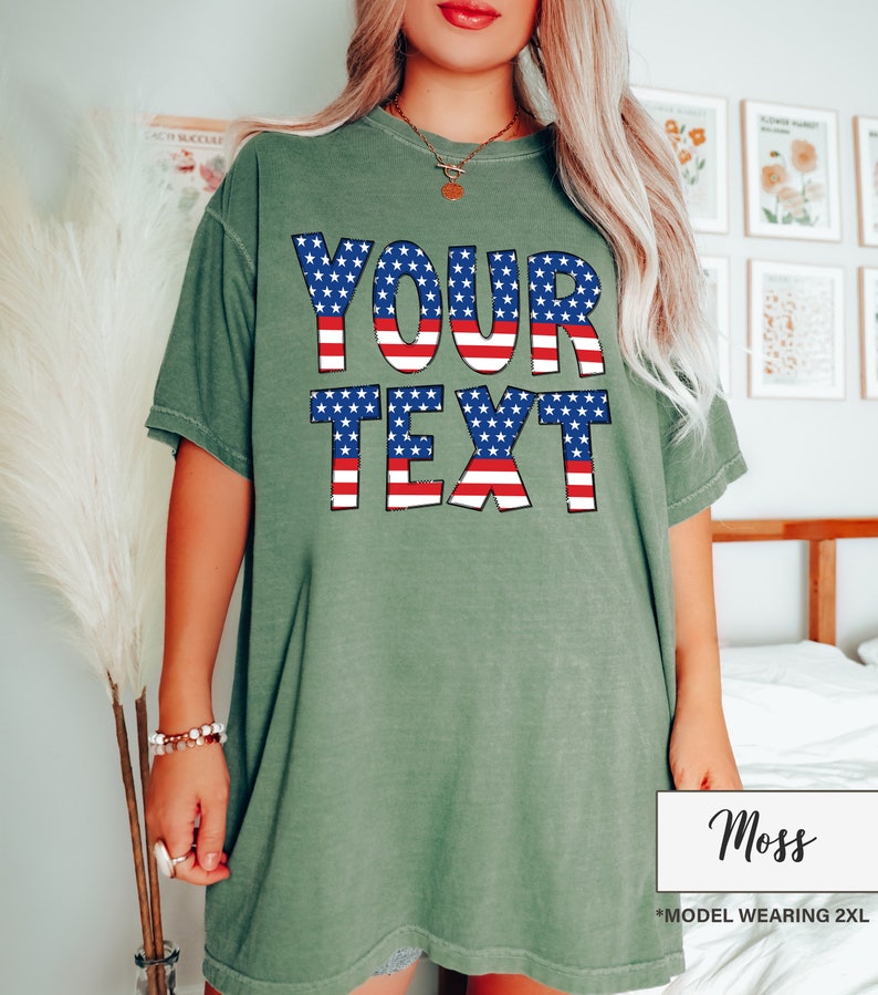 May include: Moss green t-shirt with the words "YOUR TEXT" in a patriotic design of red and white stripes and blue stars. The shirt is a loose fit and has short sleeves. The model is wearing a 2XL size.