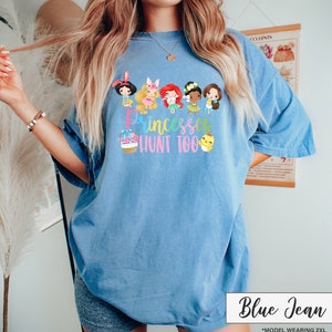 May include: Blue jean colored t-shirt with a graphic of cartoon princesses and the text "Princesses Hunt Too". The shirt is a light blue color and has a relaxed fit. The text "Blue Jean" is in a white box at the bottom.