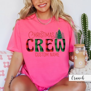Może przedstawiać: Jasnor&oacute;żowa koszulka z napisem "Christmas Crew" w czerwono-zieloną kratę. Koszulka zawiera r&oacute;wnież grafikę choinki i napis "Custom Name". Widoczny jest r&oacute;wnież szklany kubek z wzorem stokrotki.