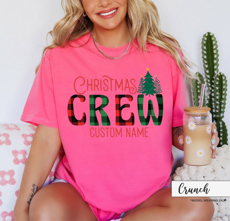 Może przedstawiać: Jasnor&oacute;żowa koszulka z napisem "Christmas Crew" w czerwono-zieloną kratę. Koszulka zawiera r&oacute;wnież grafikę choinki i napis "Custom Name". Widoczny jest r&oacute;wnież szklany kubek z wzorem stokrotki.