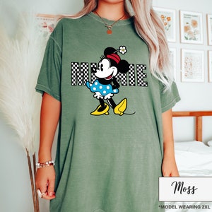 May include: Moss green t-shirt featuring a Minnie Mouse graphic. The graphic includes the word "MINNIE" in a black and white checkered pattern. Minnie Mouse is depicted in her classic outfit with a blue polka dot dress and yellow shoes.