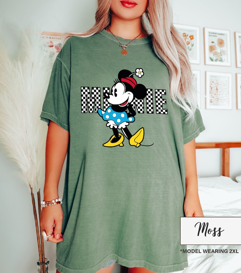 May include: Moss green t-shirt featuring a Minnie Mouse graphic. The graphic includes the word "MINNIE" in a black and white checkered pattern. Minnie Mouse is depicted in her classic outfit with a blue polka dot dress and yellow shoes.