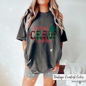 Może przedstawiać: Ciemnoszara koszulka z napisem "CHRISTMAS CREW" w czerwono-zieloną kratę. Koszulka posiada r&oacute;wnież zieloną grafikę choinki oraz napis "CUSTOM NAME" w kolorze czerwonym. Koszulka jest w kolorze vintage comfort.