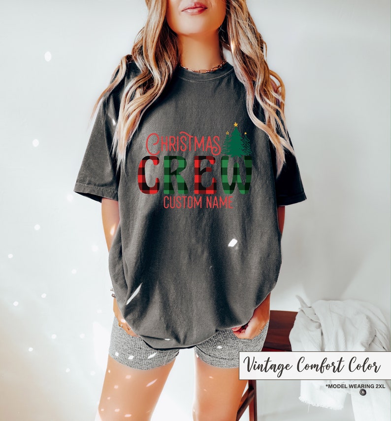 Może przedstawiać: Ciemnoszara koszulka z napisem "CHRISTMAS CREW" w czerwono-zieloną kratę. Koszulka posiada r&oacute;wnież zieloną grafikę choinki oraz napis "CUSTOM NAME" w kolorze czerwonym. Koszulka jest w kolorze vintage comfort.