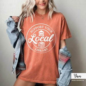 May include: Burnt orange t-shirt with a white circular graphic that reads "Support Your Local Library". The graphic includes a building and a stack of books. The model is wearing a denim jacket.
