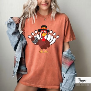 May include: Burnt orange t-shirt featuring a cartoon turkey wearing a pilgrim hat and holding a bowling ball. The turkey is surrounded by bowling pins. The shirt is a comfortable, casual style.
