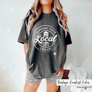 May include: Dark gray t-shirt with a white circular graphic that reads "Support Your Local Library" with a house and book illustration. The shirt is a vintage comfort color and the model is wearing a 2XL.