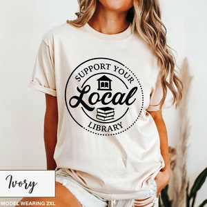May include: Ivory-colored t-shirt with a black circular graphic that reads "Support Your Local Library." The graphic includes a stylized library building and a stack of books. The shirt is a short-sleeved, casual style.