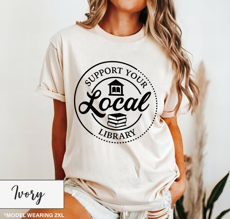 May include: Ivory-colored t-shirt with a black circular graphic that reads "Support Your Local Library." The graphic includes a stylized library building and a stack of books. The shirt is a short-sleeved, casual style.
