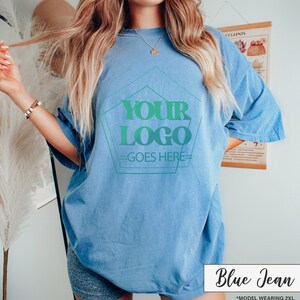 May include: A light blue, oversized t-shirt with the text "YOUR LOGO GOES HERE" in a green gradient design. The shirt is labeled "Blue Jean" in a white box. The model is wearing a 2XL size.