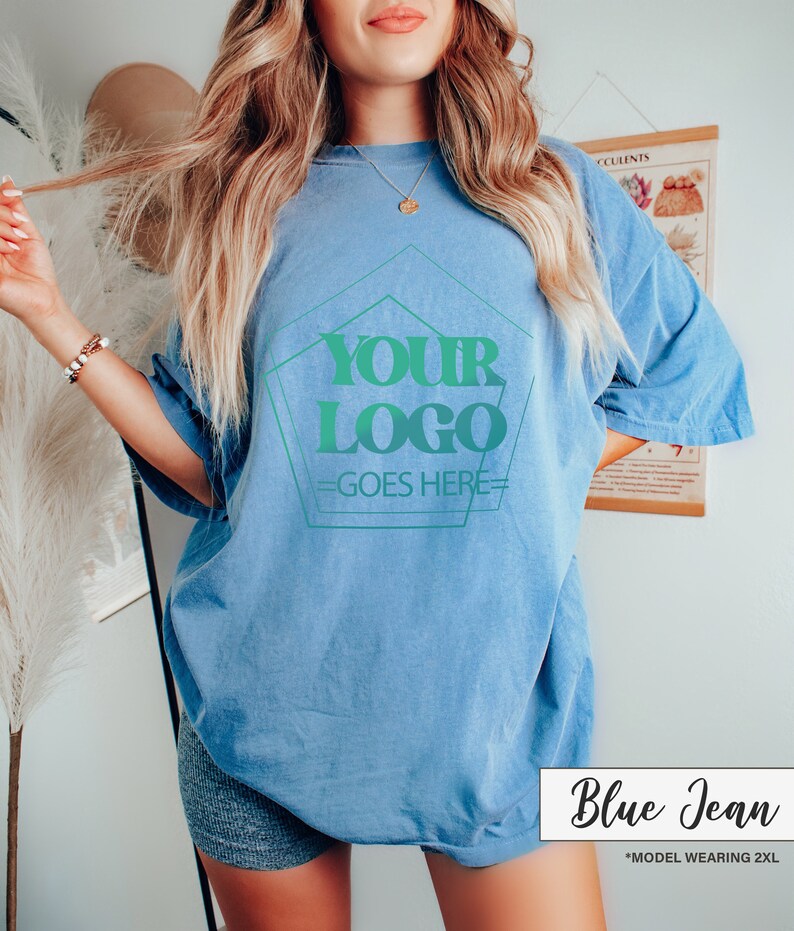 May include: A light blue, oversized t-shirt with the text "YOUR LOGO GOES HERE" in a green gradient design. The shirt is labeled "Blue Jean" in a white box. The model is wearing a 2XL size.