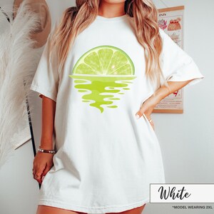 May include: White oversized t-shirt with a graphic of a lime wedge in the water. The text "White" is written in black at the bottom of the image. The text "*MODEL WEARING 2XL" is written in black below the word "White".