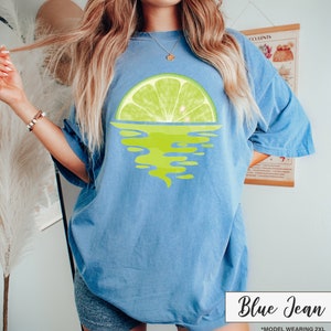 May include: A blue jean t-shirt with a lime wedge graphic, the lime wedge is half submerged in water, the water is depicted as a green swirl. The text "Blue Jean" is in white text on a black background. The text "*MODEL WEARING 2XL" is in white text on a black background.
