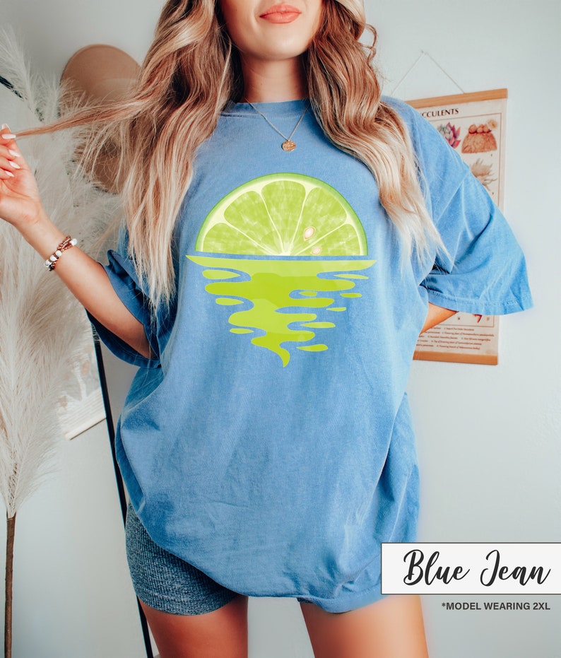 May include: A blue jean t-shirt with a lime wedge graphic, the lime wedge is half submerged in water, the water is depicted as a green swirl. The text "Blue Jean" is in white text on a black background. The text "*MODEL WEARING 2XL" is in white text on a black background.